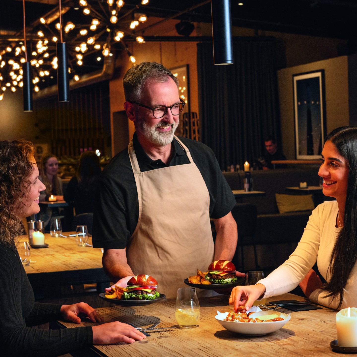 A waiter serves pretzel burgers to guests in a warmly lit restaurant.