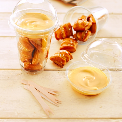 Plastic cup with Pretzel Bites and cheese dip along with wooden forks on wooden surface.