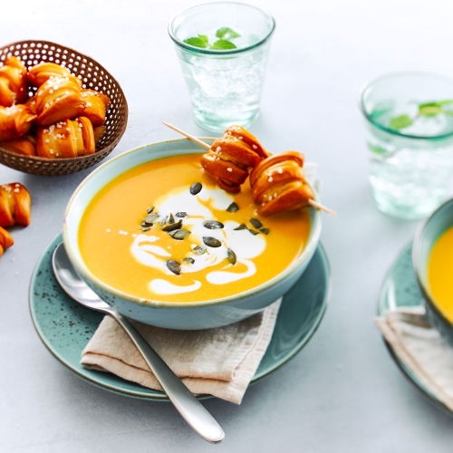 Zwei Teller Kuerbissuppe mit Ditsch Pretzel Bites auf Holzspiessen und Kuerbiskernen.