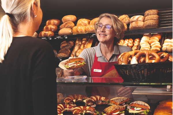 Ditsch-Baeckerei mit modernen Verkaufstheke. Eine Frau hinter der Theke übergibt ein belegtes Laugengebäck an eine weitere Frau.