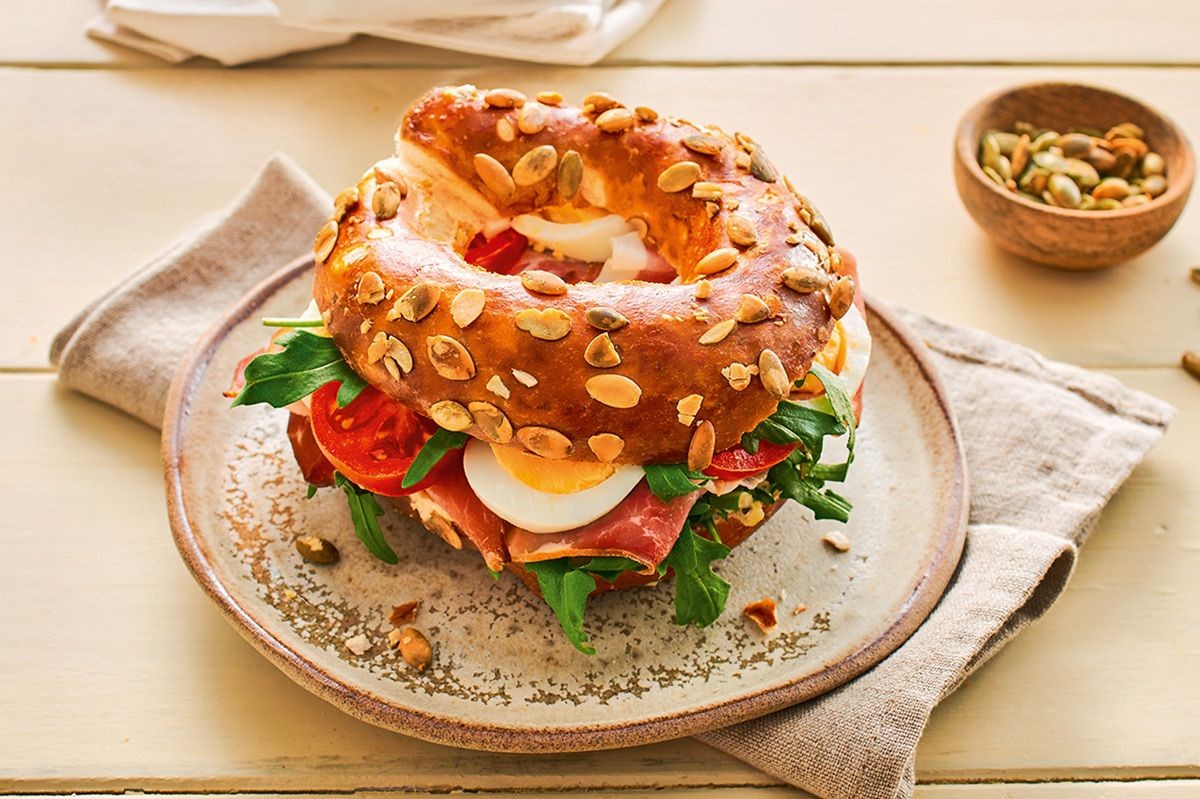 Belegter Laugenkringel mit Kürbiskernen, Rucola, Tomaten, Ei und Schinken auf einem Teller.
