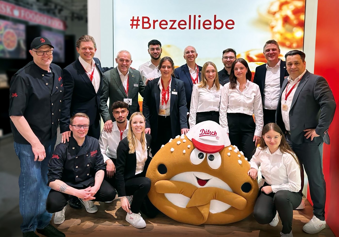 A group of employees standing in front of an exhibition booth, with a pretzel-shaped mascot in the center.