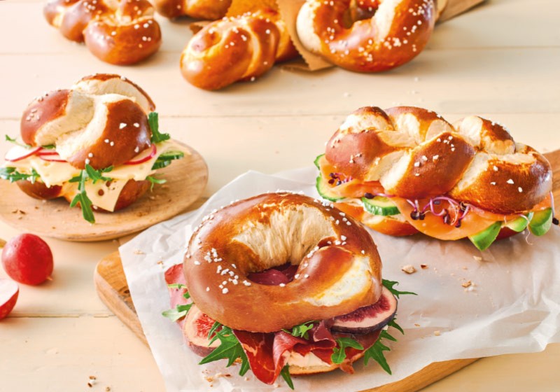 Assorted pretzel rolls and sandwiches filled with cheese, cured meat, vegetables, and greens on a wooden table.