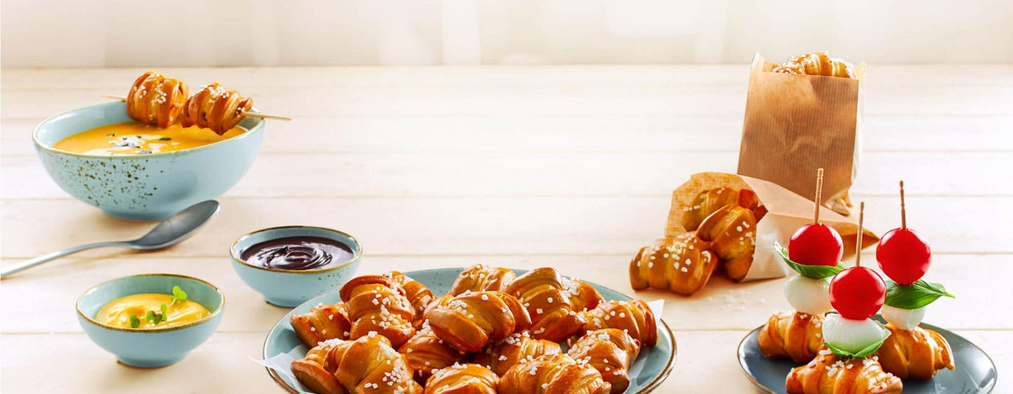 A selection of mini pretzel bites served with dipping sauces, soup, and tomato-mozzarella skewers on a bright wooden table.