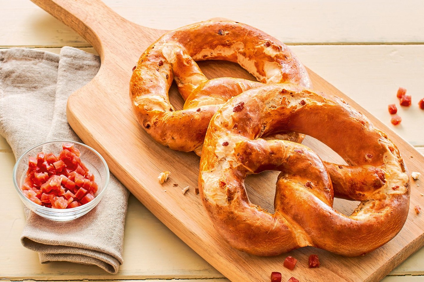 Zwei große Laugenbrezeln mit Schinkenstückchen auf einem Holzbrett, daneben eine kleine Schale mit gewürfeltem Schinken.