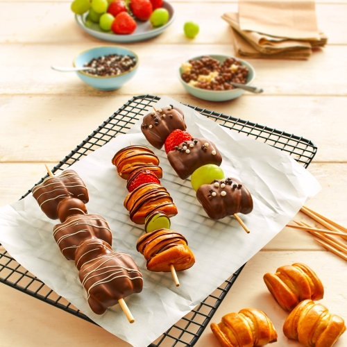 Drei schokoladenueberzogene Pretzel-Bite-Spiesse mit Beeren auf schwarzem Gitterrost.